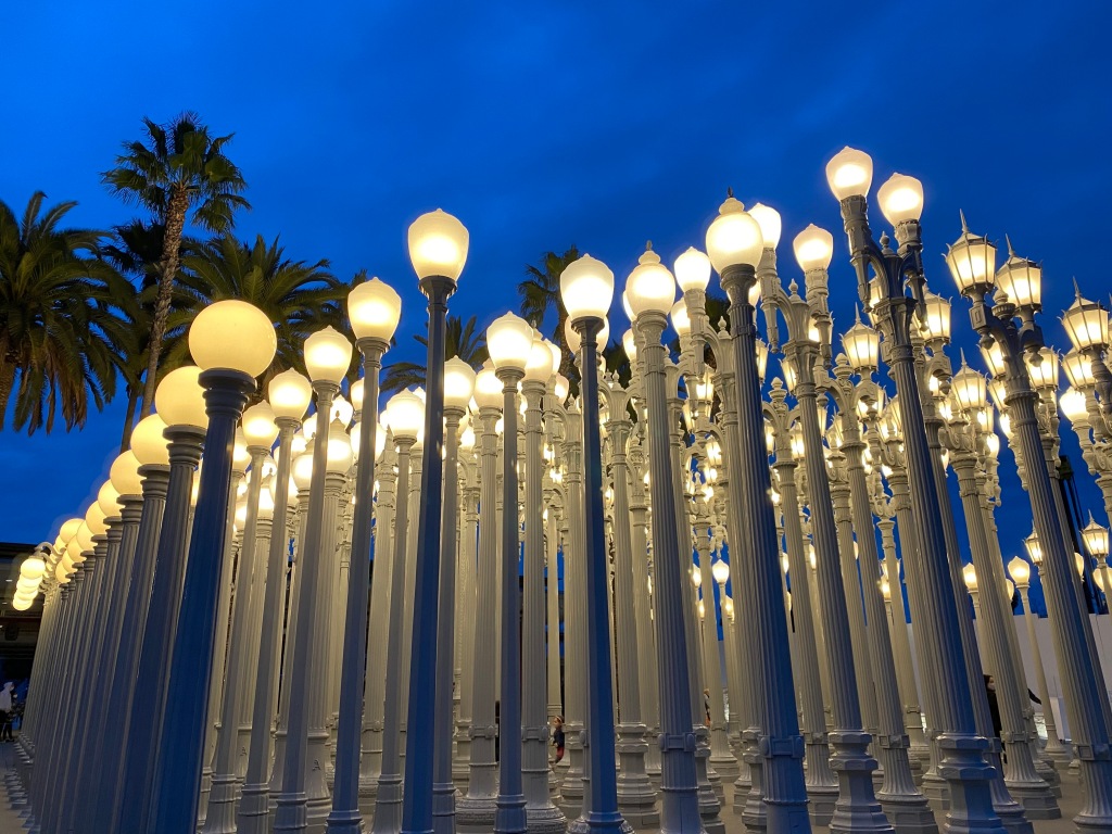 A multitude of various street lamps arranged in rows, lit lamps perched above off-white columns, stand in from of a dusky sky with palm trees in the background.