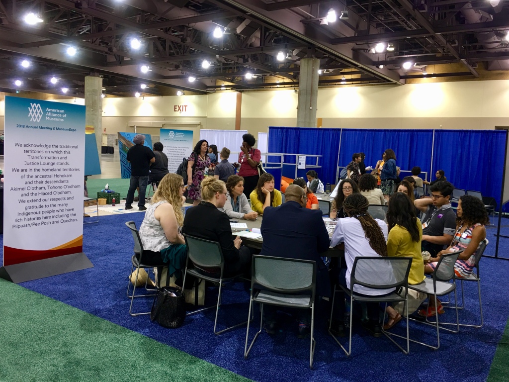 A few dozen professionals of a variety of races are seated around a table conversing with others chatting in the background. A sign with the AAM Logo that says "2018 Annual Meeting & MuseumExpo and a land acknowledgement sits to the side.